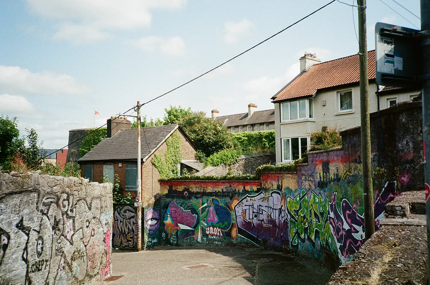 Narrow street lined with stone walls covered in colorful graffiti and houses with tiled roofs in the background under a partly cloudy sky.
