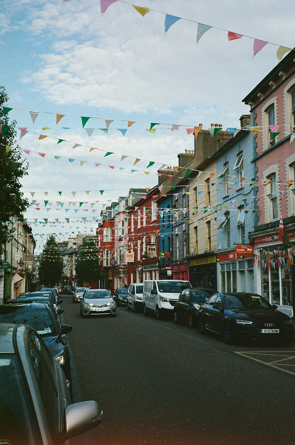 Street lined with colorful buildings and parked cars, decorated with multiple strings of multicolored triangular flags overhead under a cloudy blue sky.