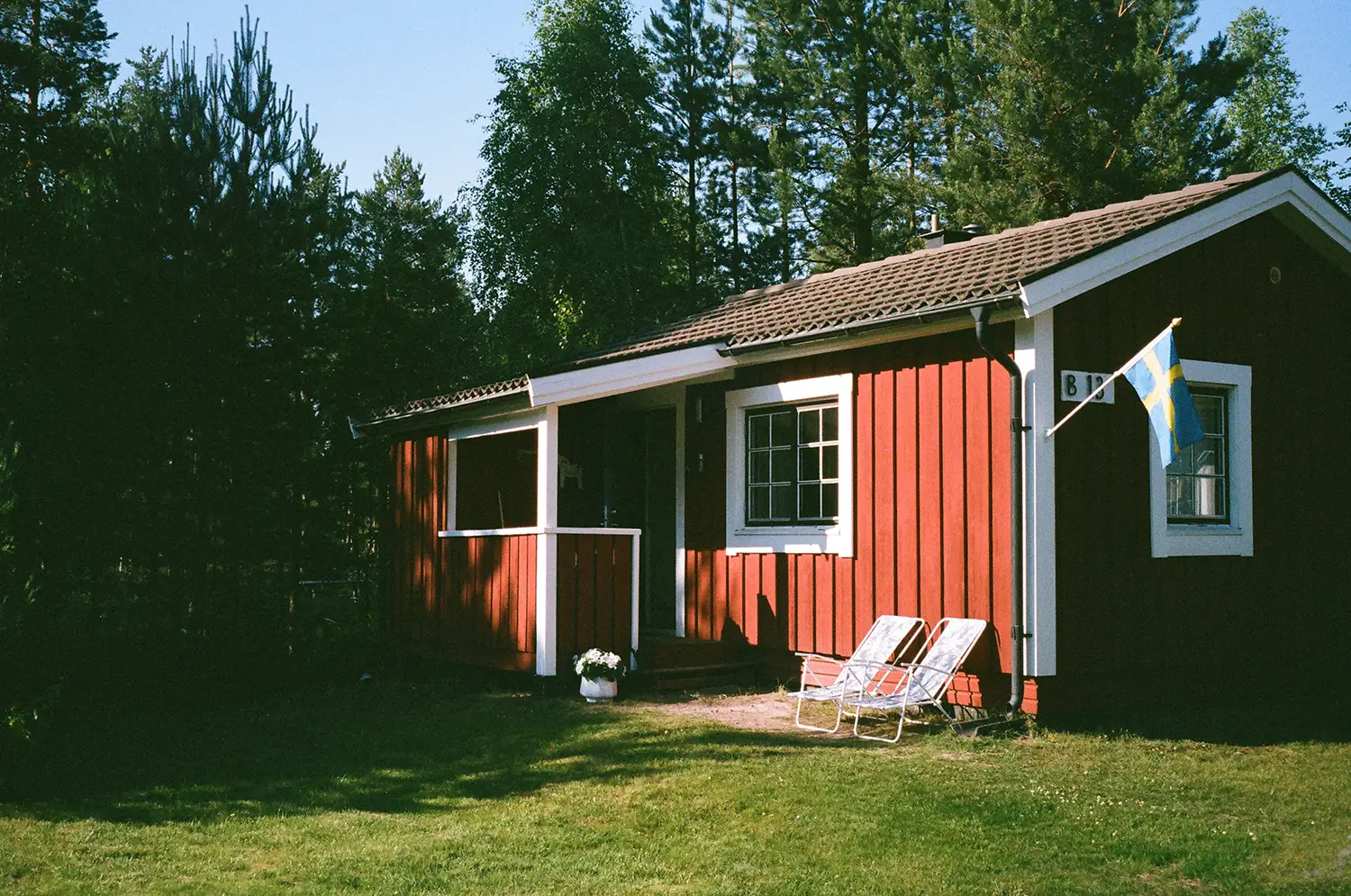 Red wooden cabin with white trim in a sunny green yard, two lawn chairs, and a Swedish flag on the wall.