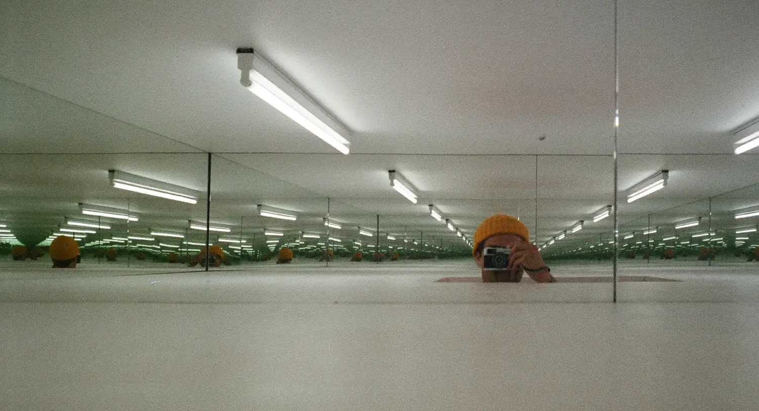 Person wearing an orange beanie taking a photo in a room with mirrors creating infinite reflections of the person and ceiling lights.