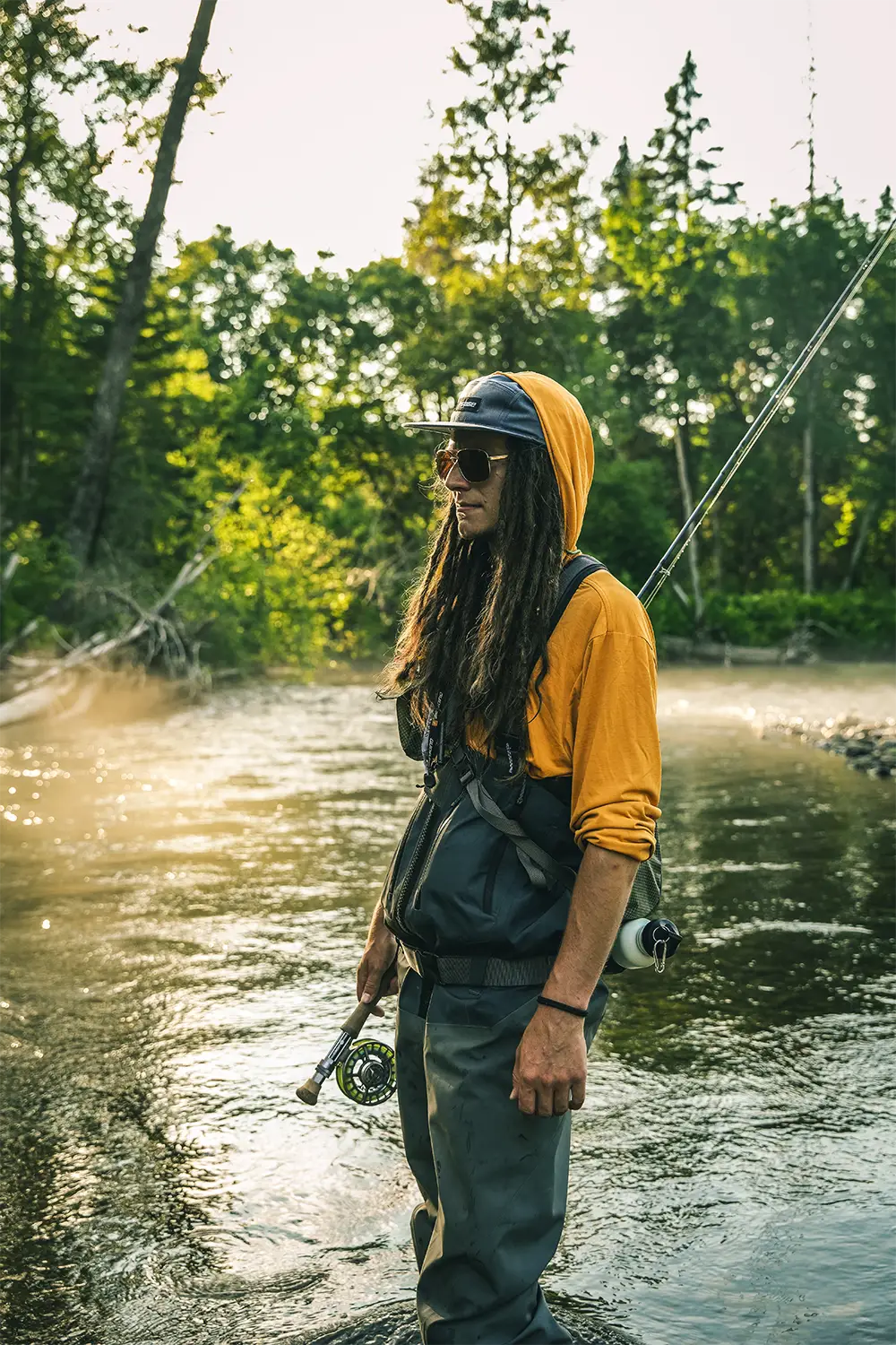 Young man with long hair and sunglasses holding a fishing rod standing in a river surrounded by trees.