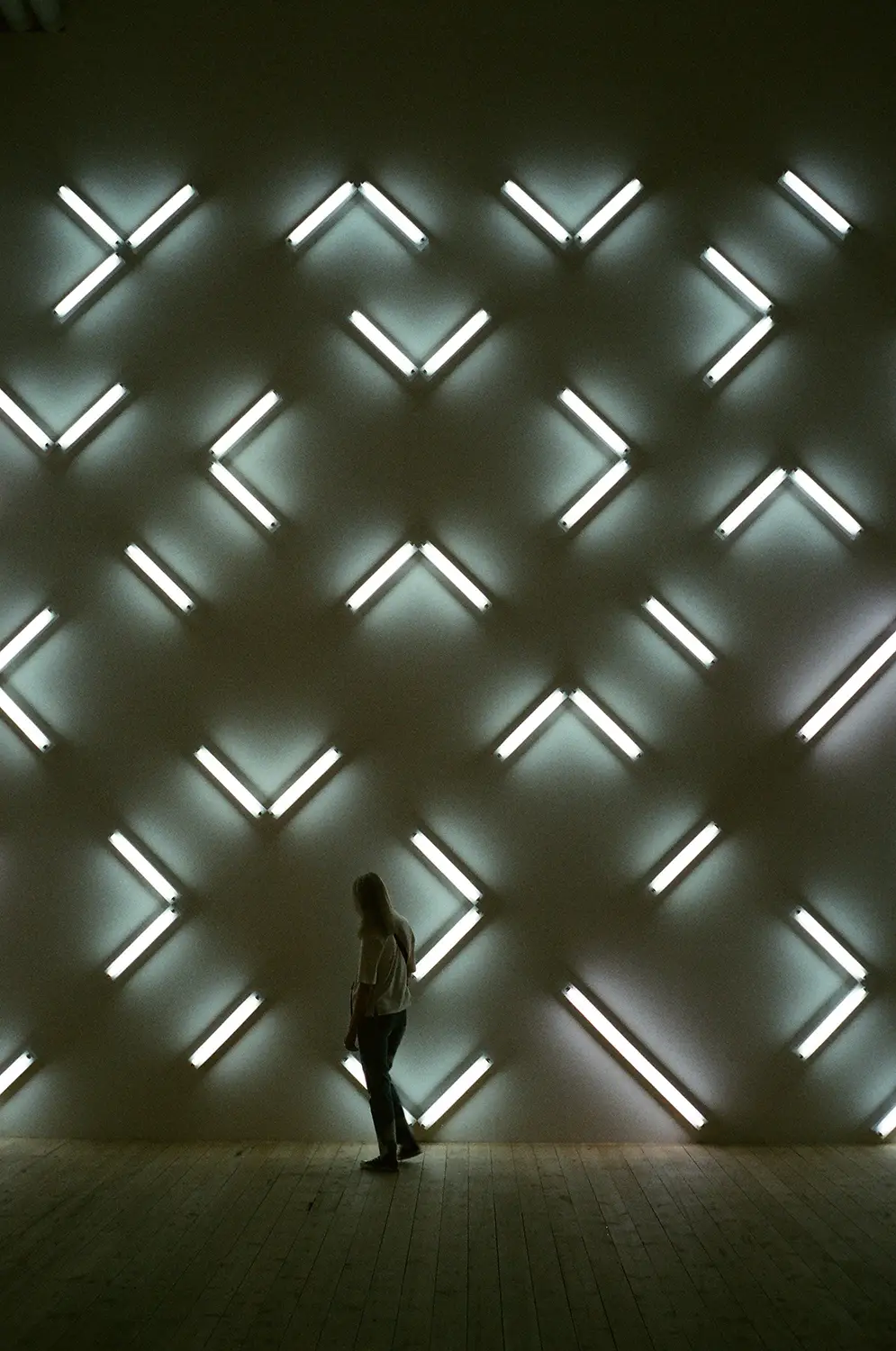 Person standing in a dark room with a wall displaying a grid of illuminated white fluorescent light tubes arranged in various angular patterns.