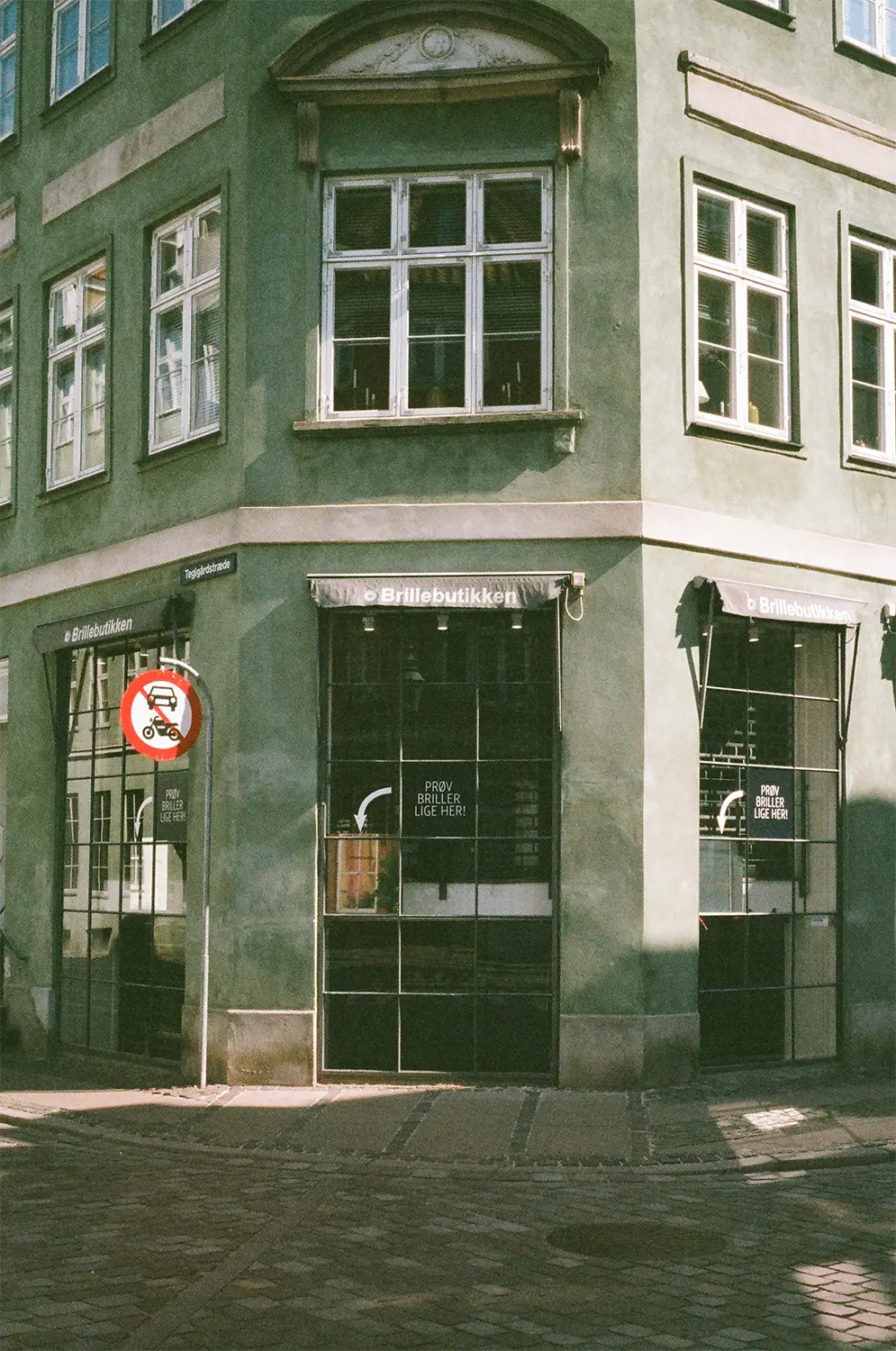 Corner of a green building with three windows above and two storefronts named Brillebutikken with signs saying 'Prøv briller lige her!'