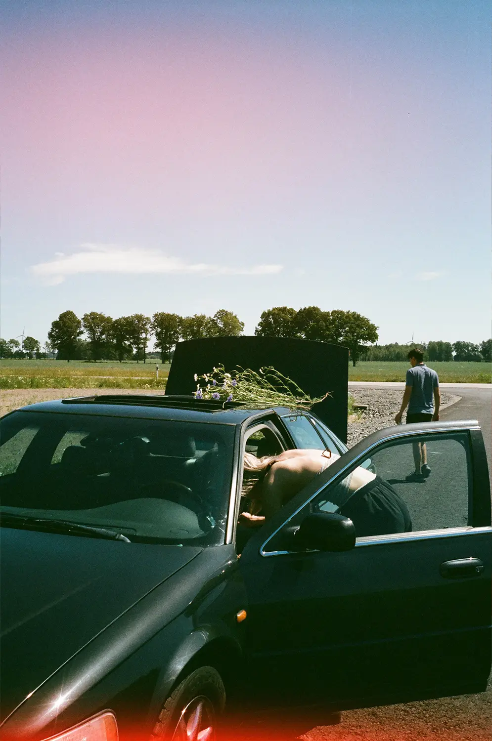 Person leaning into the open driver's side of a black car with flowers on the hood and another person walking away in the background on a rural road.