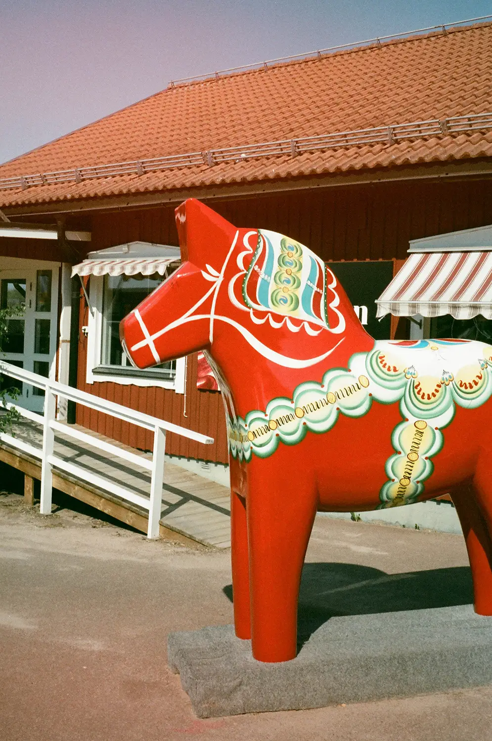 Large red Dala horse statue with traditional painted decoration in front of a building with a tiled roof and striped awnings.
