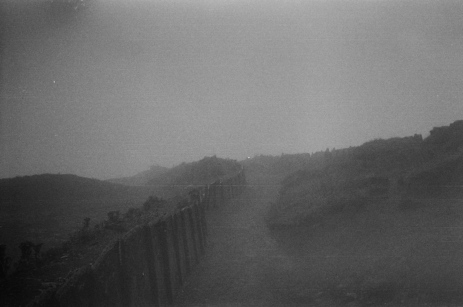 Grainy black and white image of a coastline with hills and a pier or jetty extending into the water.