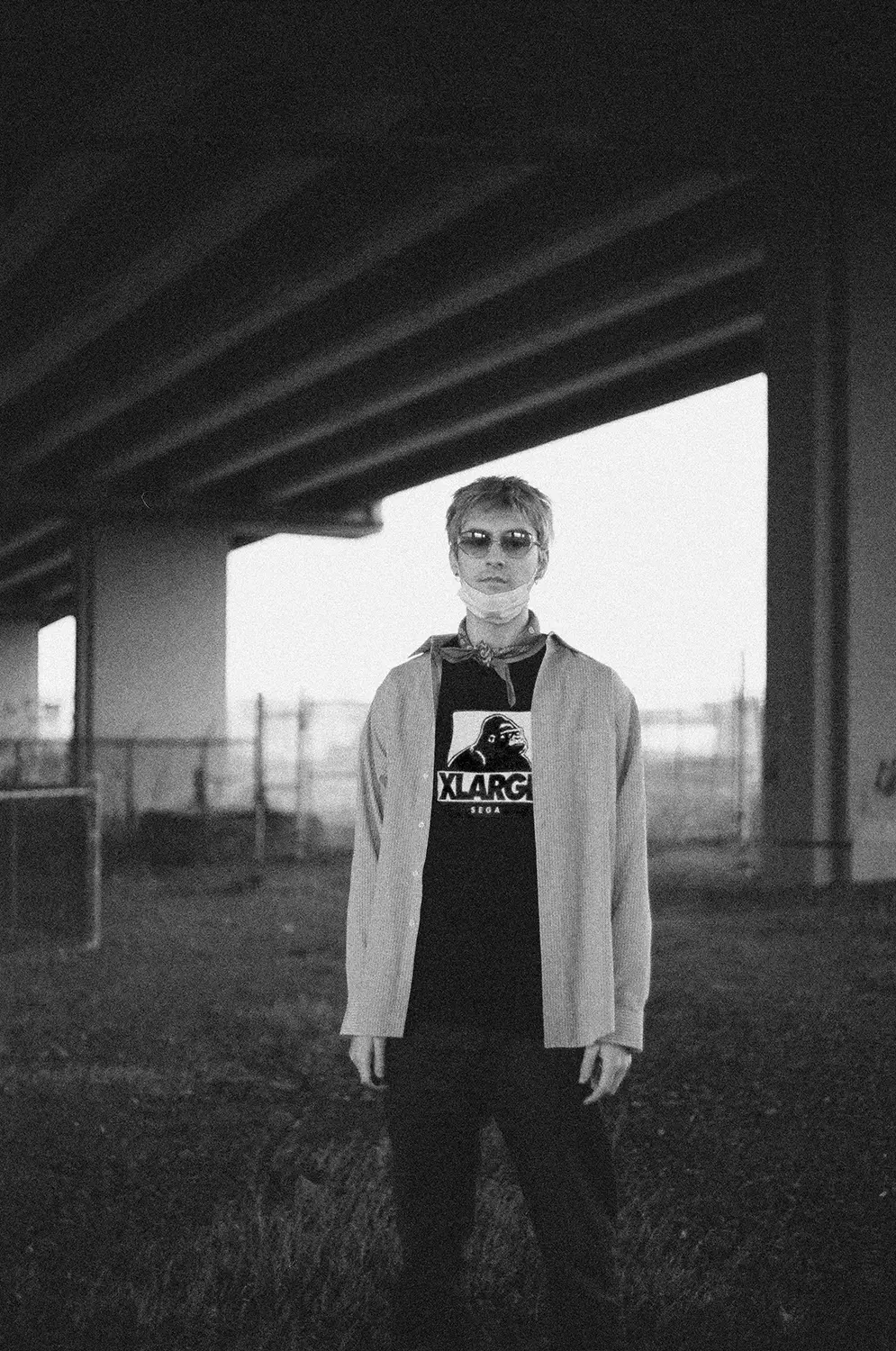 Black and white photo of a person with sunglasses and a mask under their chin standing under a bridge wearing an XLARGE t-shirt and an open shirt over it.