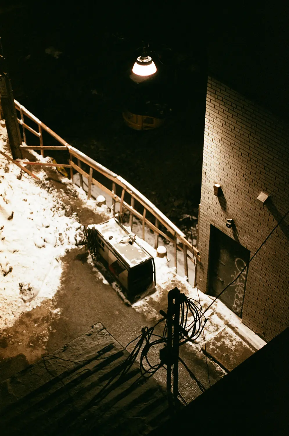 Night scene of a snow-covered alley with a wooden fence, an electrical box, and a brick wall illuminated by a single hanging street lamp.