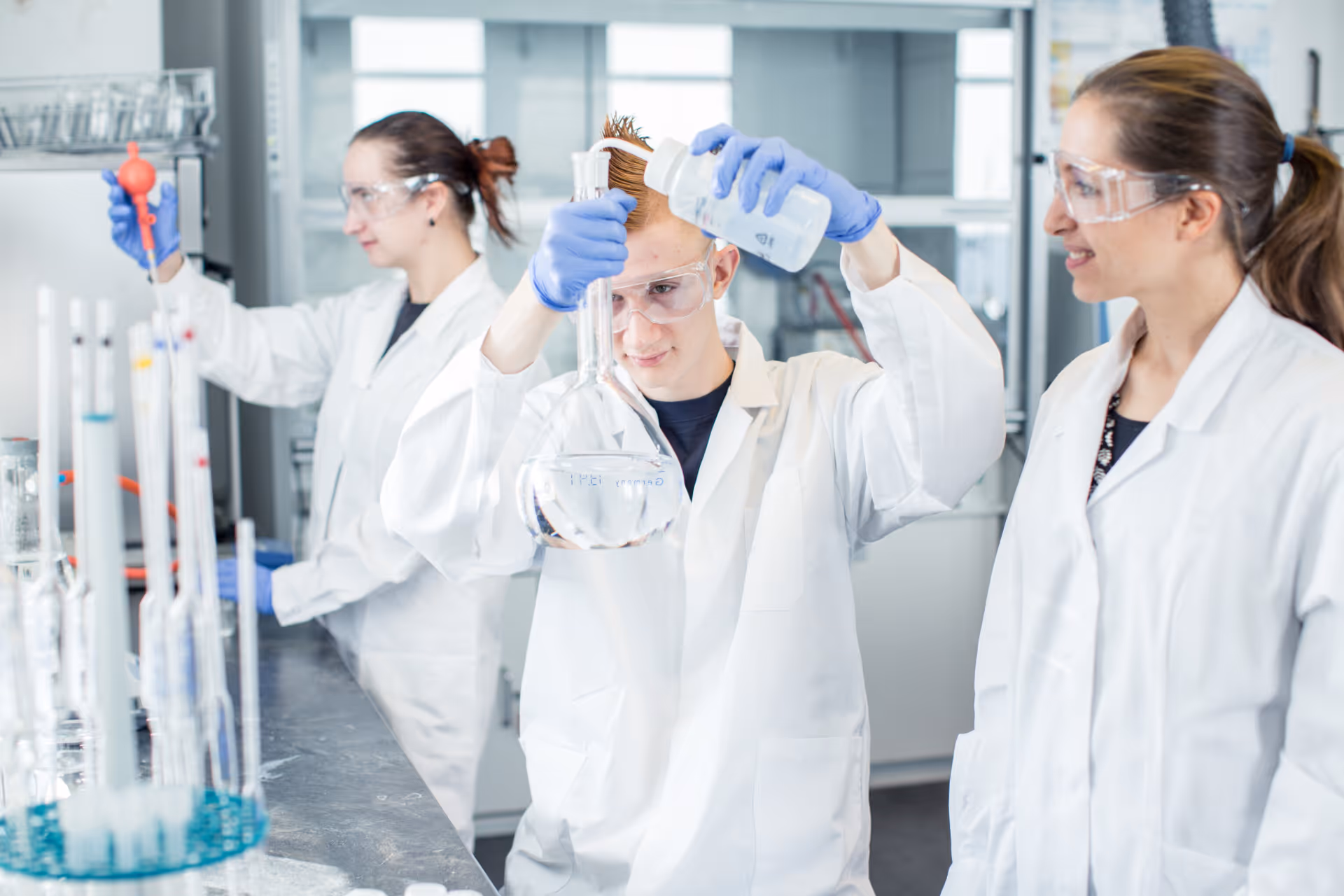 Three scientists in lab coats and safety glasses conducting experiments with clear and blue liquids in a laboratory.