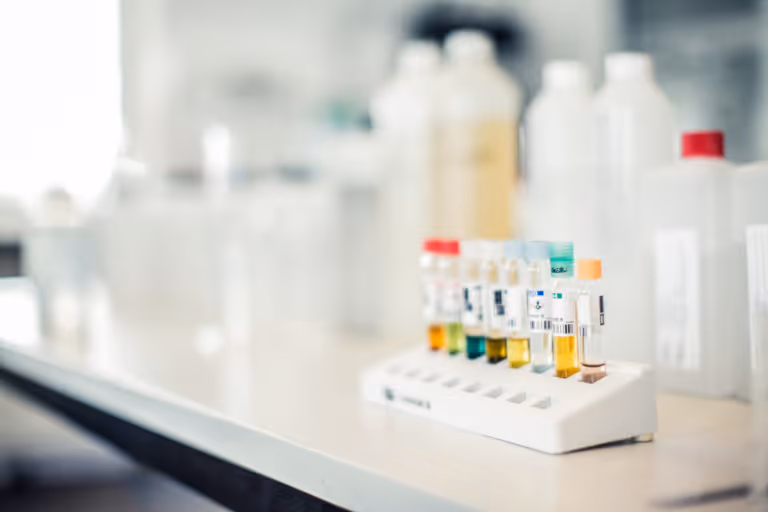 Test tubes with various colored liquids arranged in a white rack on a laboratory bench with blurred bottles in the background.