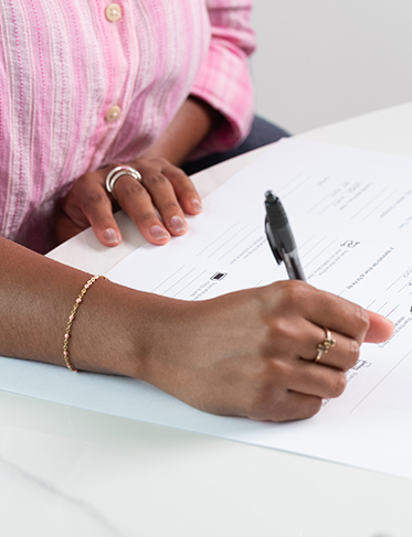 Person writing notes with a pen in an open notebook on a white table with a sticky note nearby.