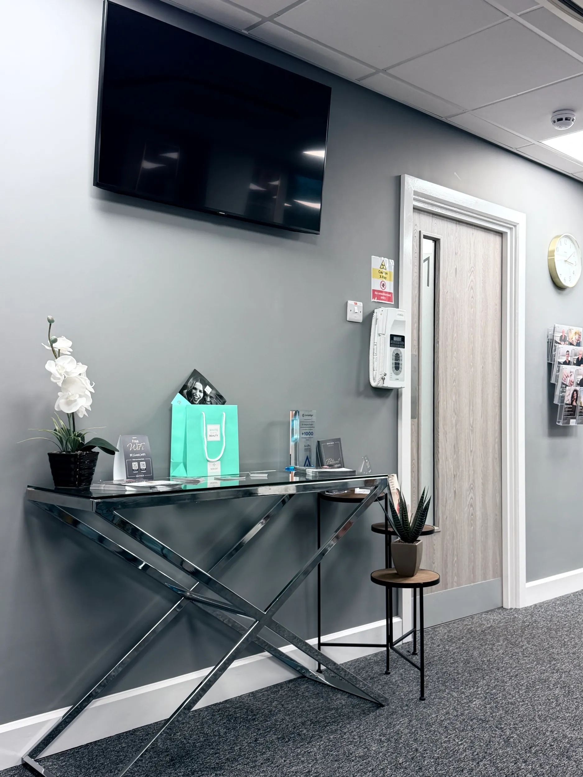Modern gray office hallway with a glass and chrome console table holding a white orchid plant, a turquoise gift bag, brochures, a wall-mounted telephone, door labeled '1', and a clock on the wall.