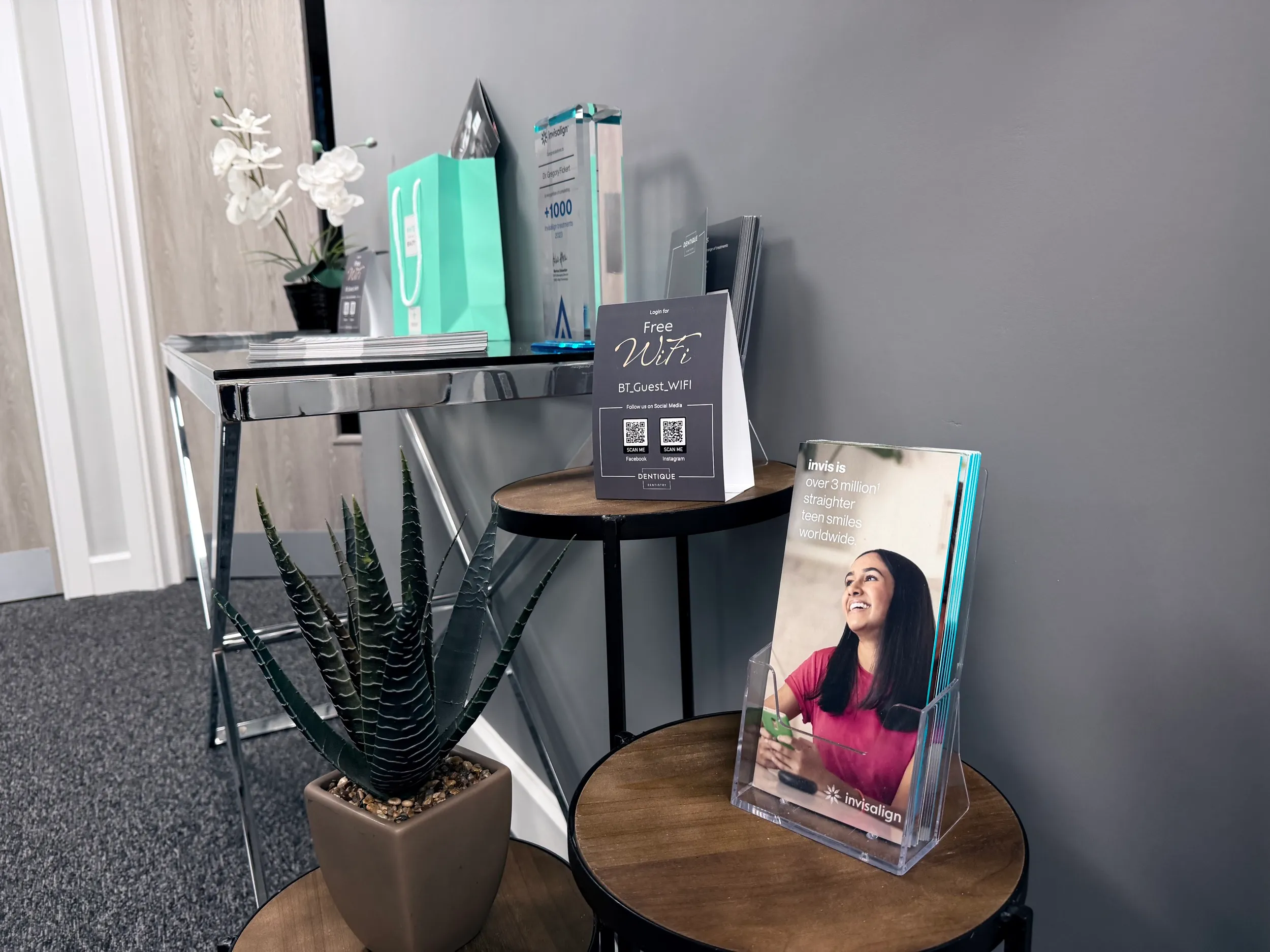 Reception area with two round wooden tables displaying an aloe plant, a free WiFi sign, and Invisalign brochures against a gray wall.