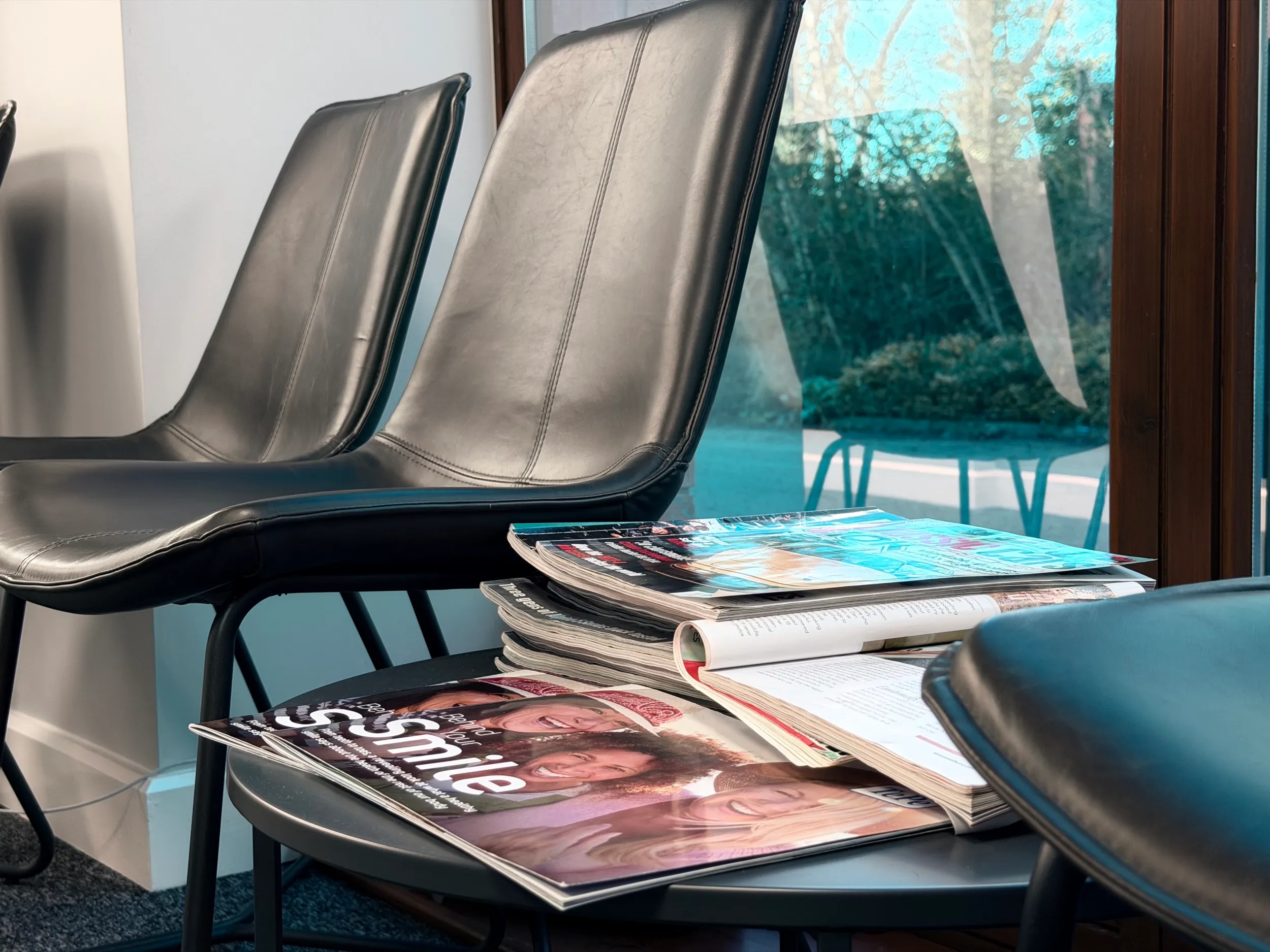 Black leather chairs around a small table stacked with magazines and newspapers in a waiting area by a large window.