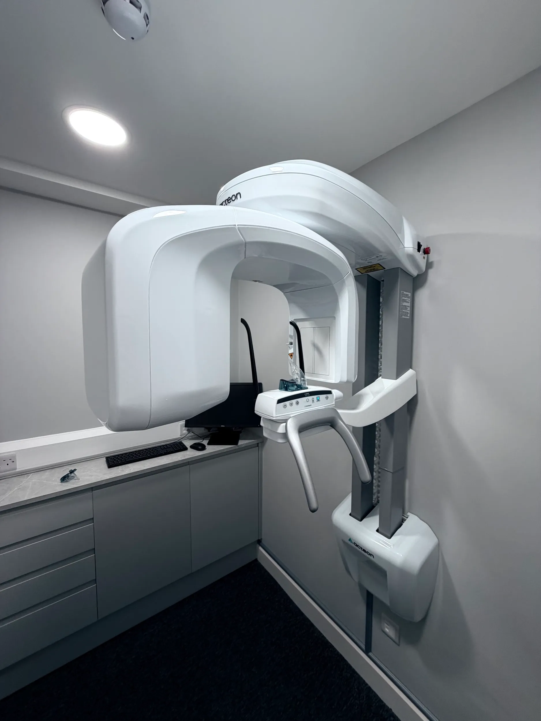 White dental panoramic X-ray machine mounted on wall in a modern examination room with gray cabinetry and computer on countertop.