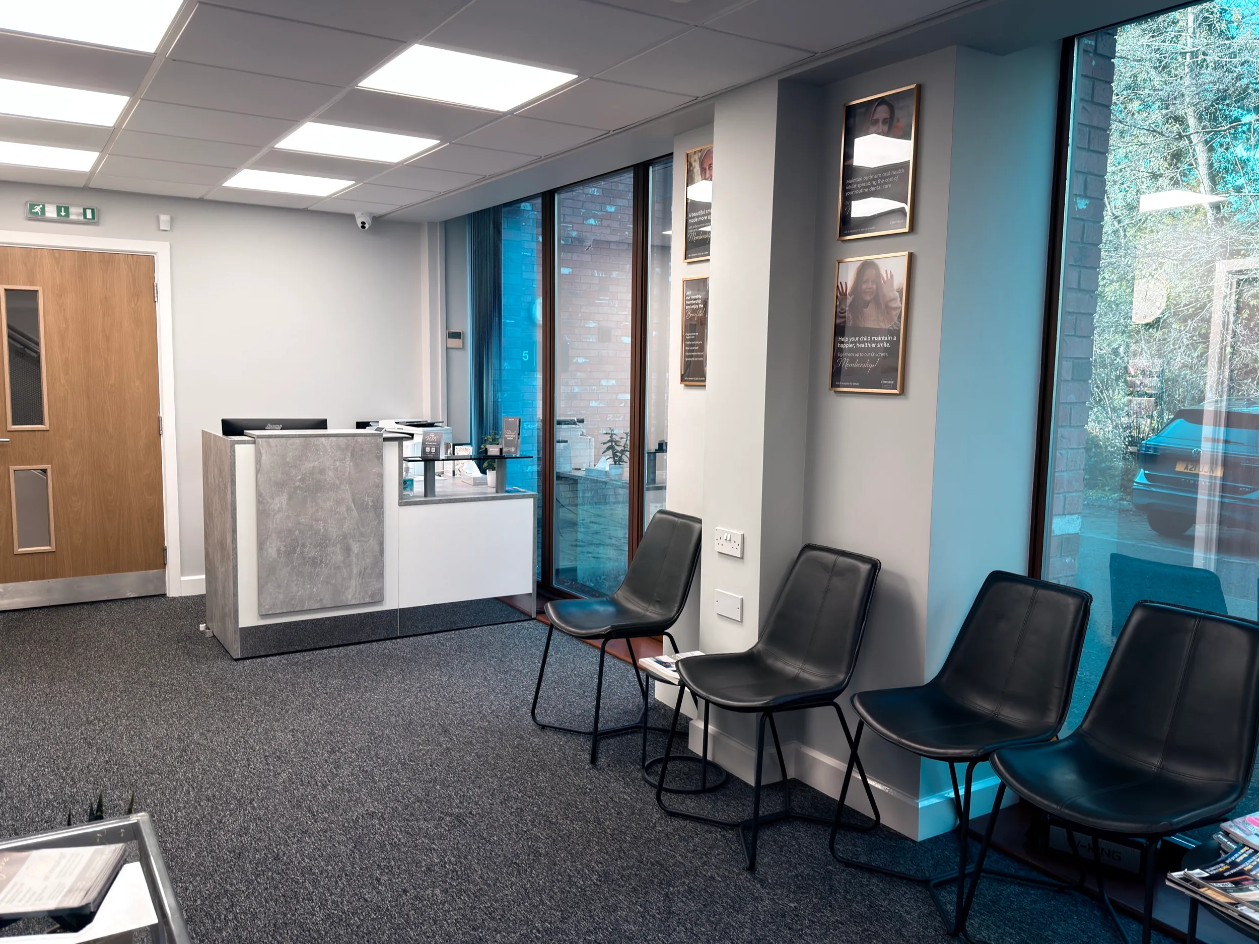 Modern reception area with a gray carpet, four black chairs against a windowed wall, and a small front desk.