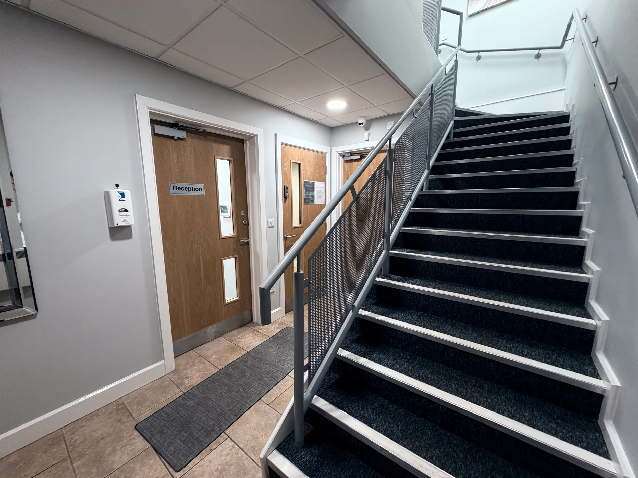 Indoor stairway with dark carpet and metal handrails next to beige tiled floor and three wooden doors, one labeled Reception.