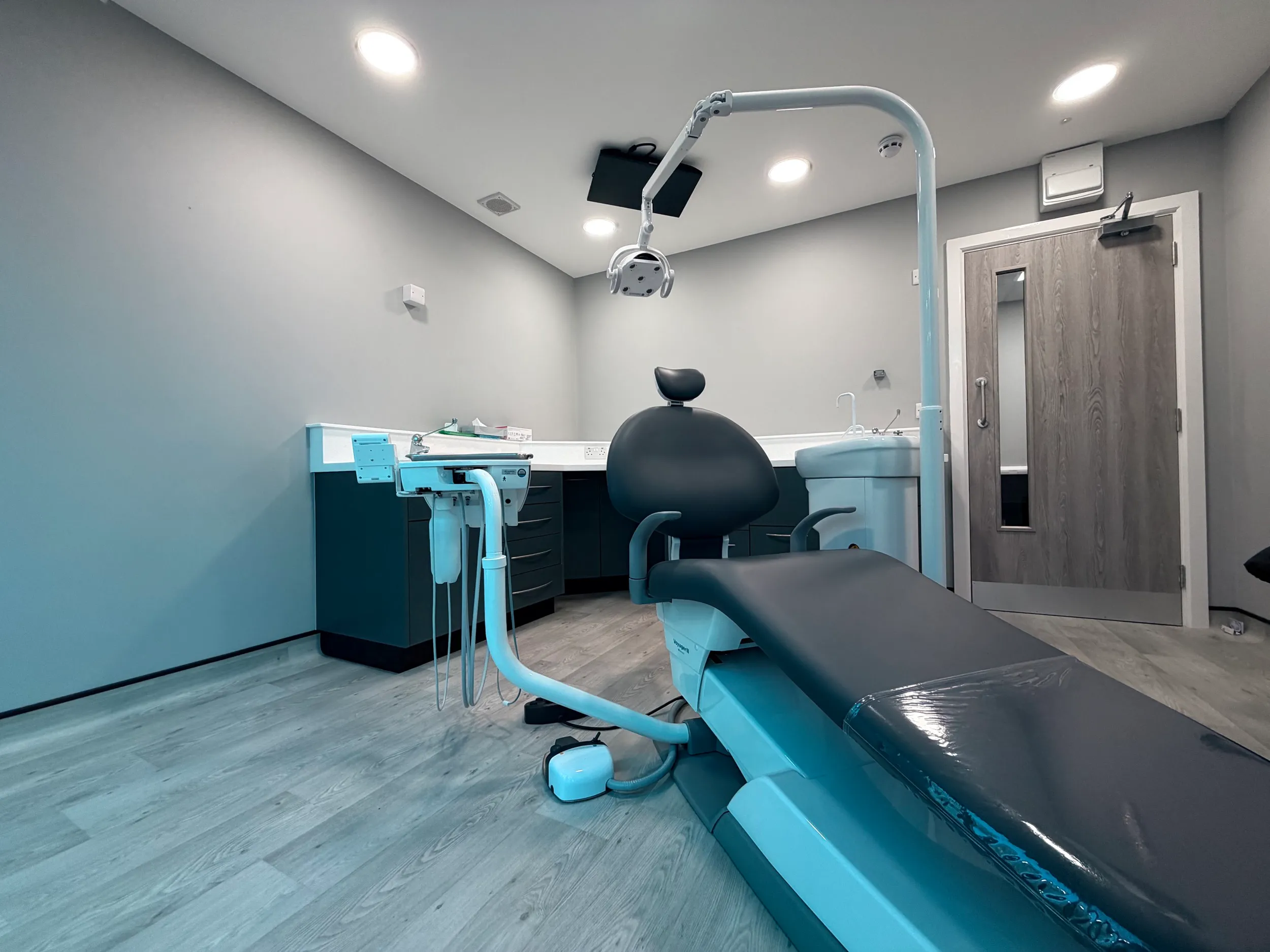 Modern dental examination room with reclining dentist chair, overhead light, and cabinetry against gray walls.