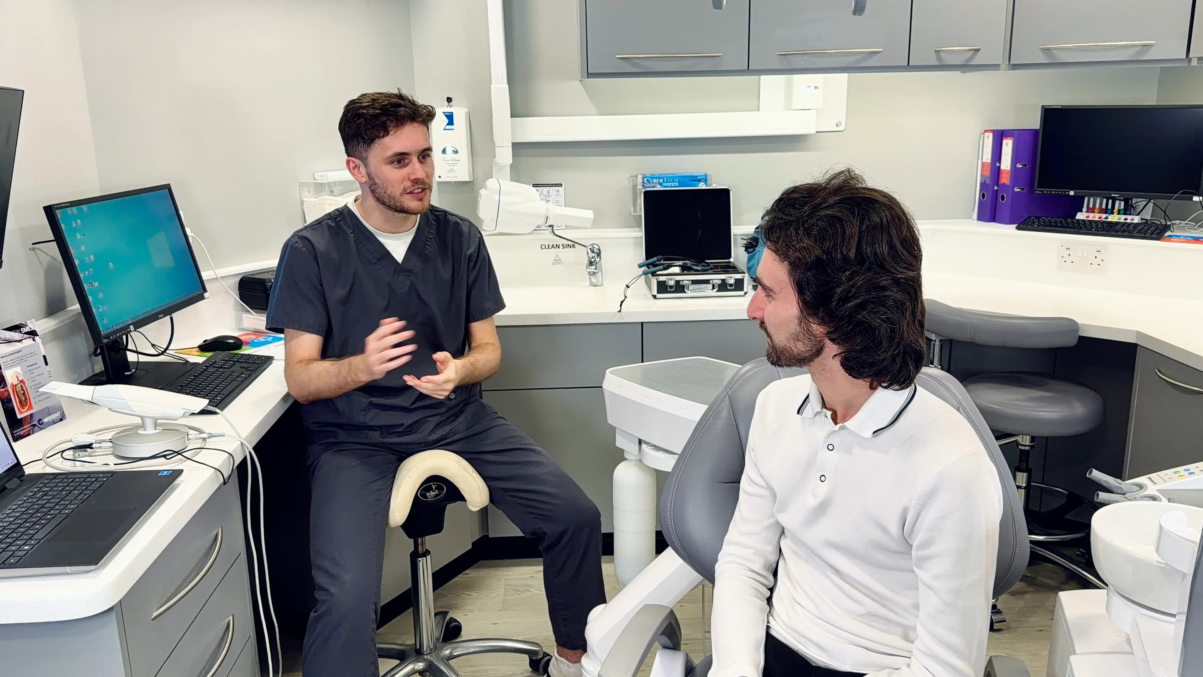 Dentist in scrubs talking to a patient seated in a dental chair in a clinic room.