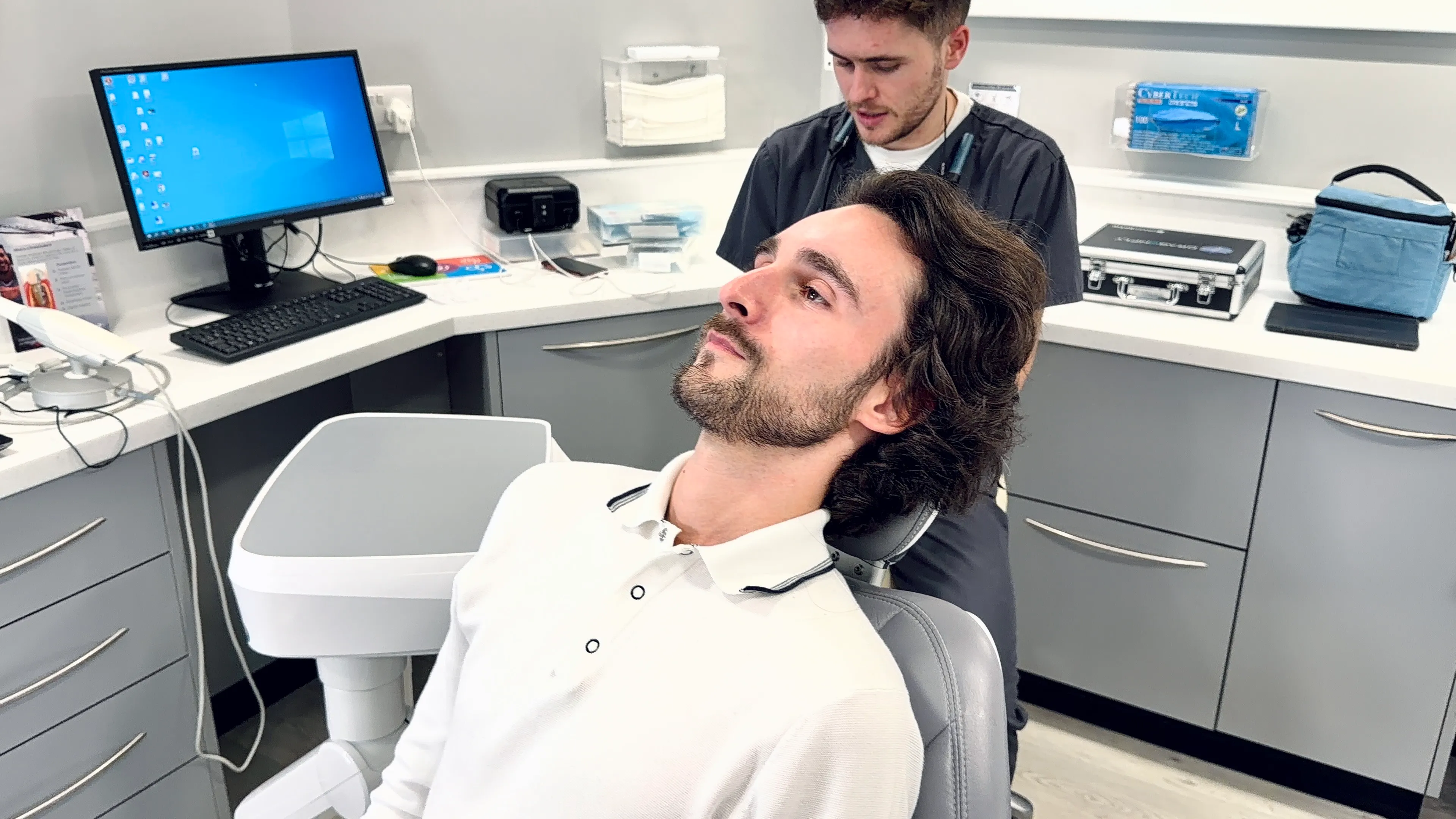 A man with dark hair and a beard reclines in a dental chair while a dental professional prepares behind him in a clinical setting.