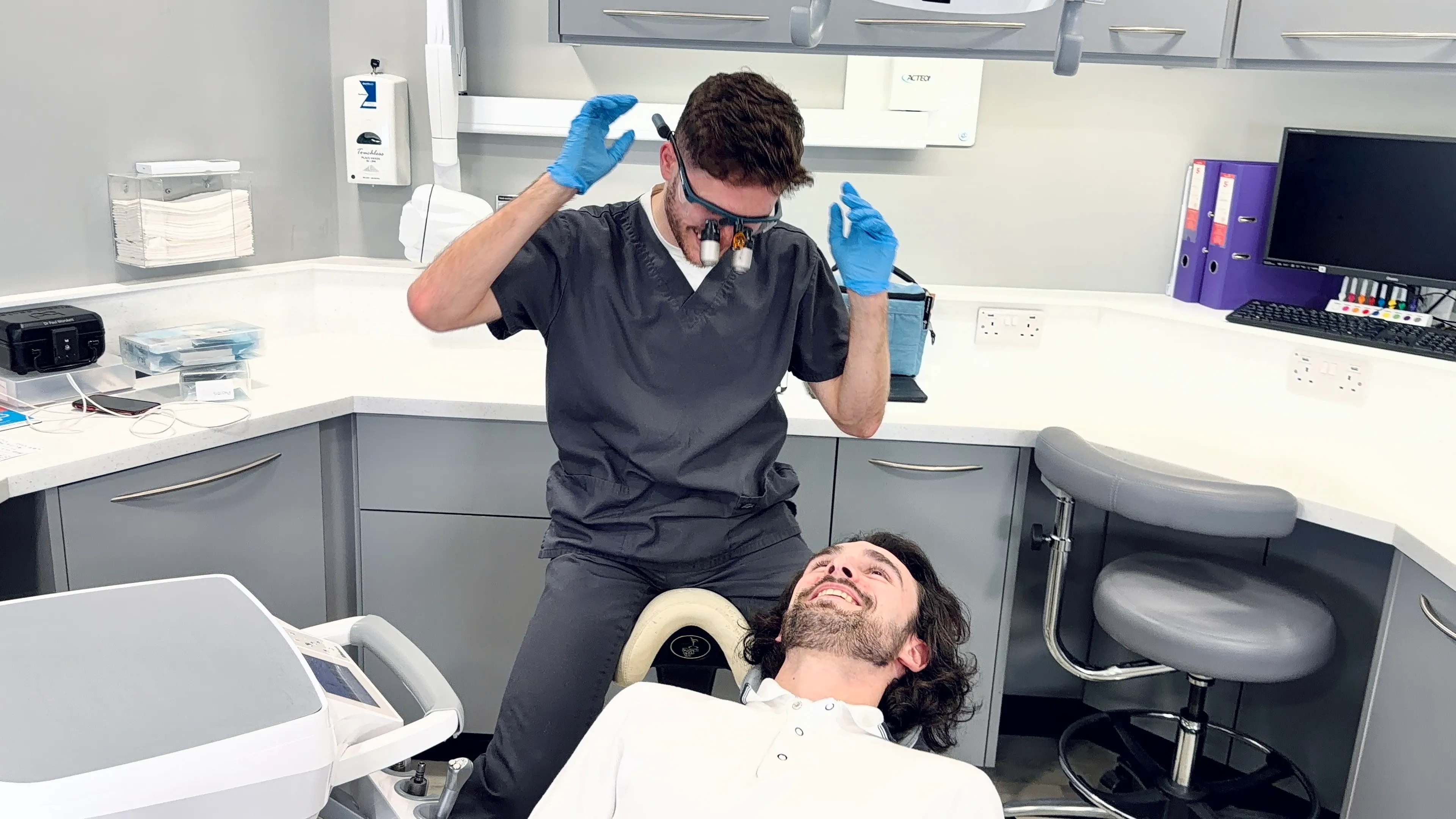 Dentist wearing magnifying glasses and blue gloves leaning over a smiling male patient in a dental clinic.