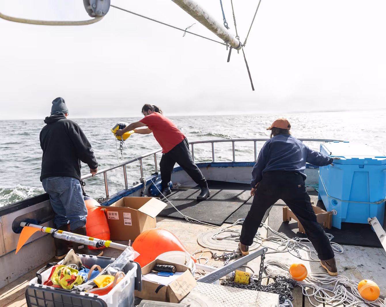 Three people working on a boat deck handling equipment near the railing on a cloudy day at sea.