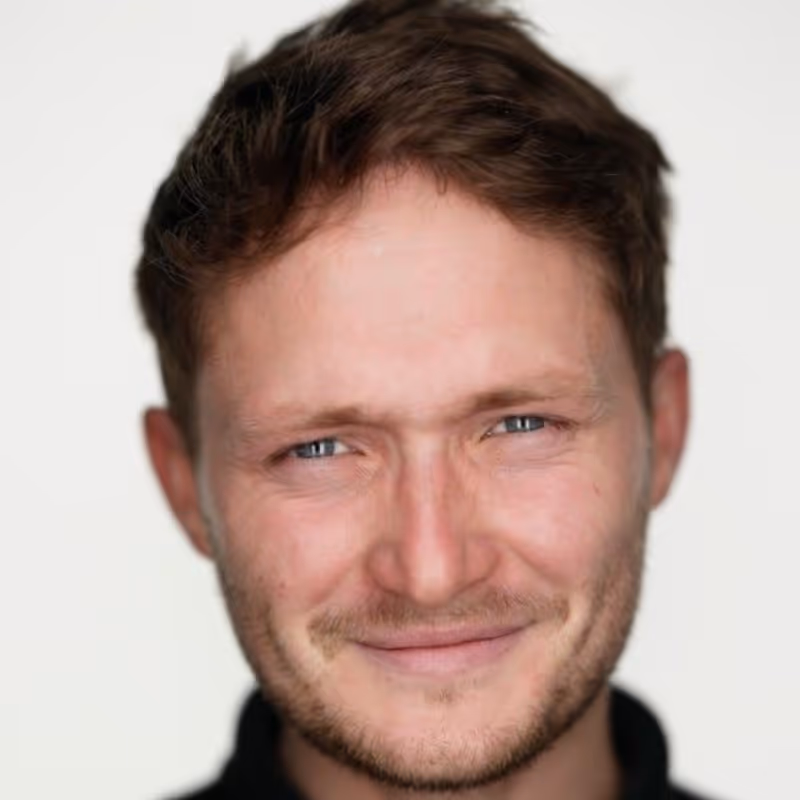 Close-up portrait of a smiling man with short brown hair and blue eyes against a plain white background.