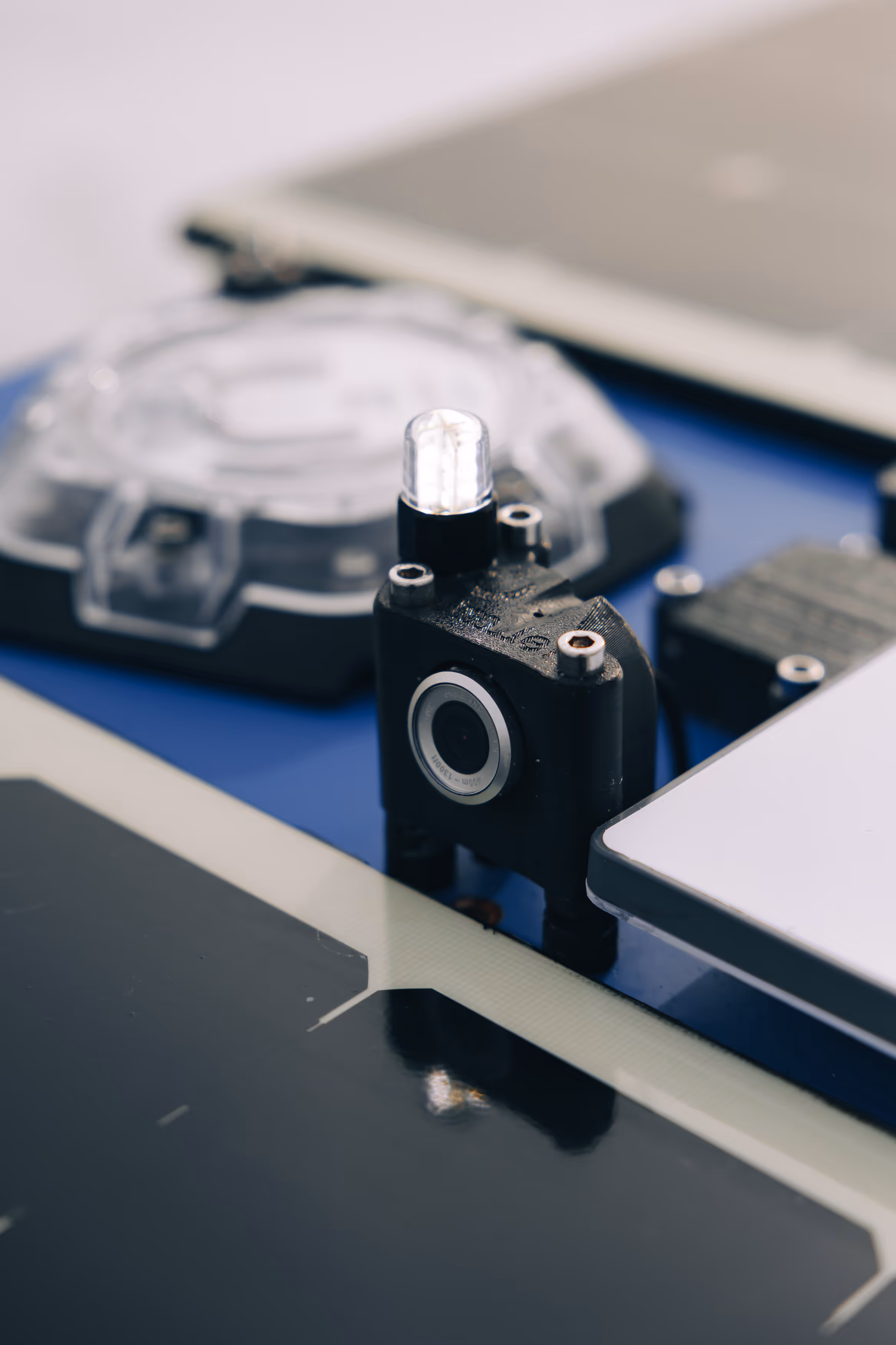 Close-up of a small black surveillance camera with an LED light on top, placed on a blue surface with other electronic devices around.
