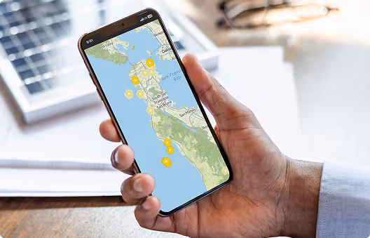 Hand holding a smartphone displaying a map of the San Francisco Bay Area with yellow icons marking locations.