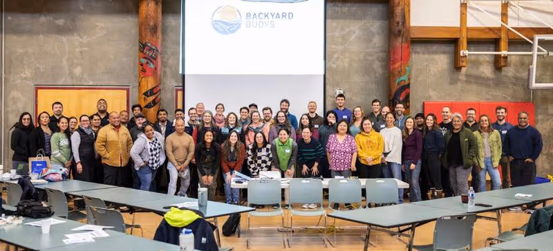 A group of people smiling in front of a projector reading "Backyard Buoys"