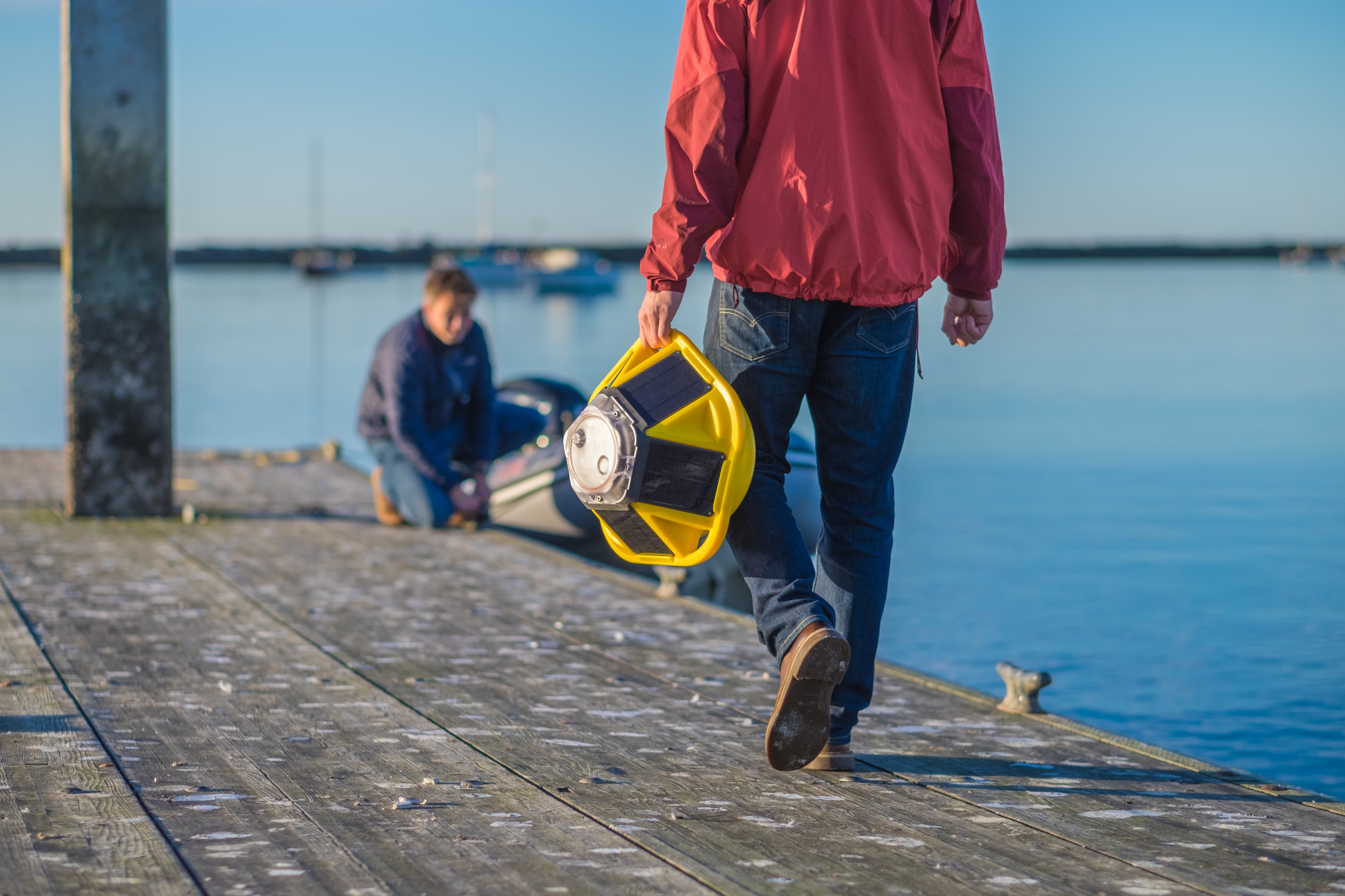 Field technician carrying Spotter ocean buoy for hand deployment from coastal dock with harbor infrastructure in background