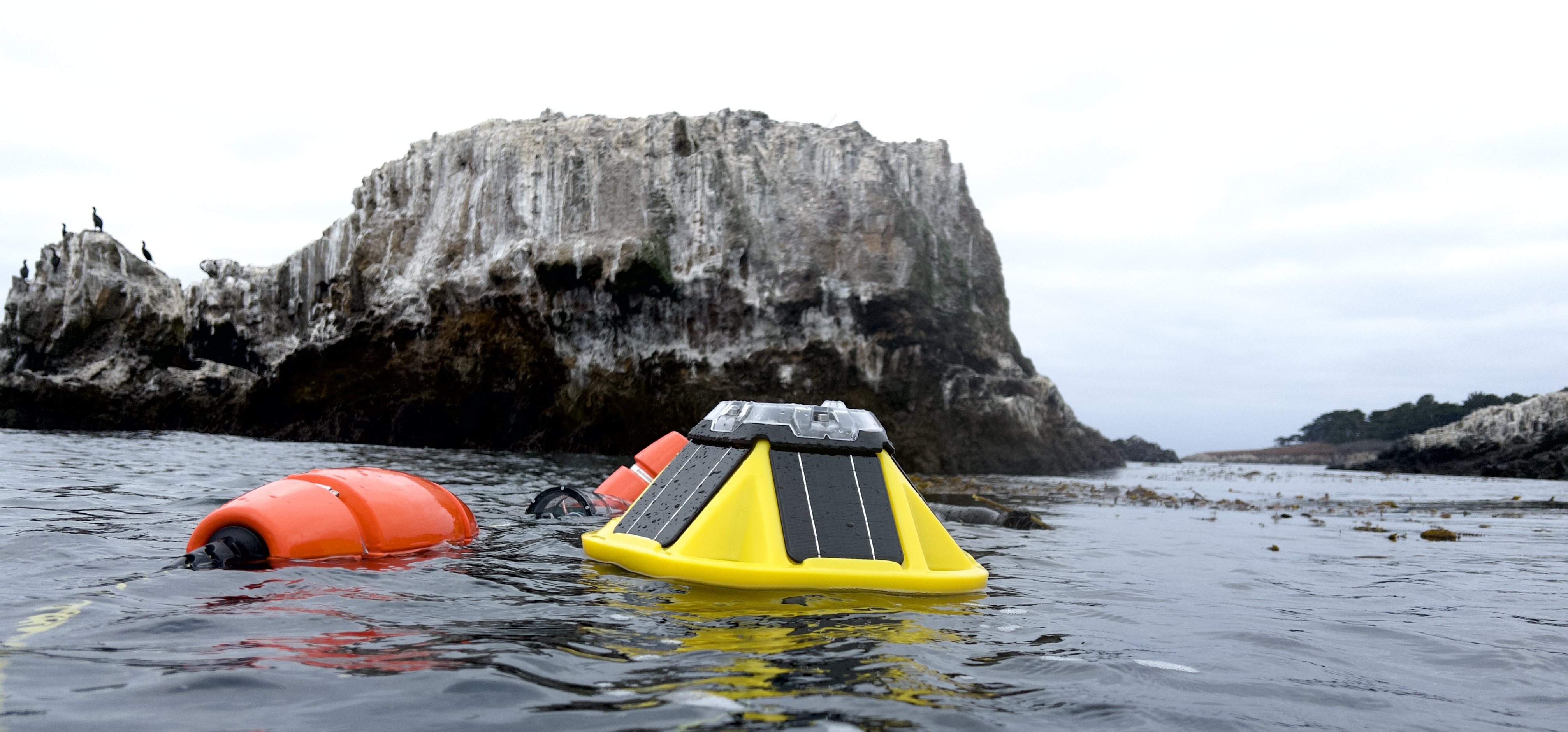 Yellow Spotter buoy and orange mooring float deployed in coastal waters near rocky island formation