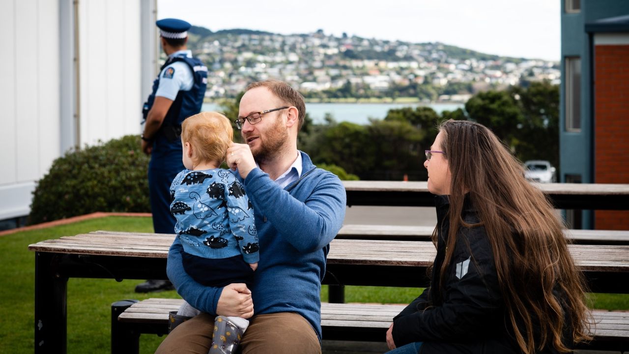 A man holds his young child at a table outside. A woman is seated talking to them. Behind is a police officer leaving.