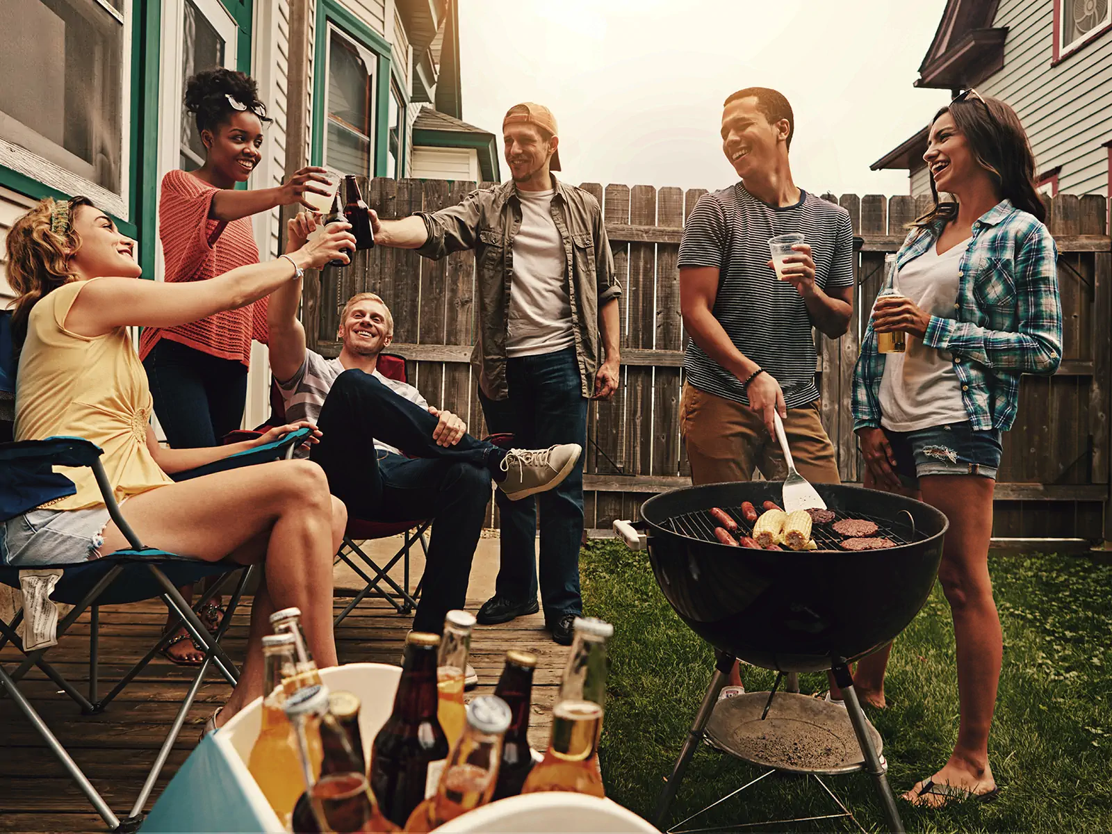 A group of friends smile and cheers as they barbecue in the backyard in front of a wooden fence