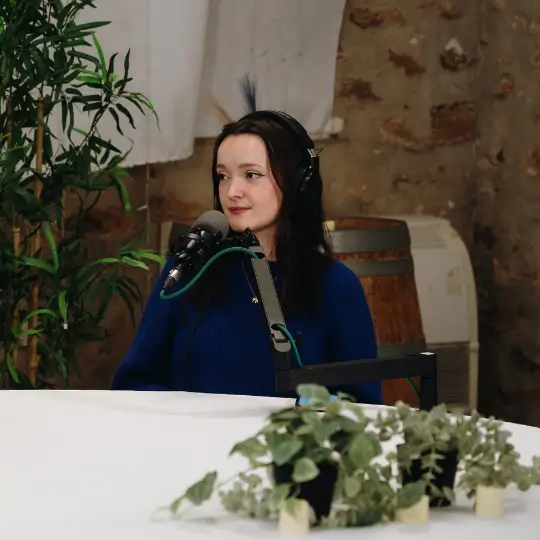 A woman wearing headphones speaking into a microphone at a table with green plants in the foreground and a brick wall in the background.