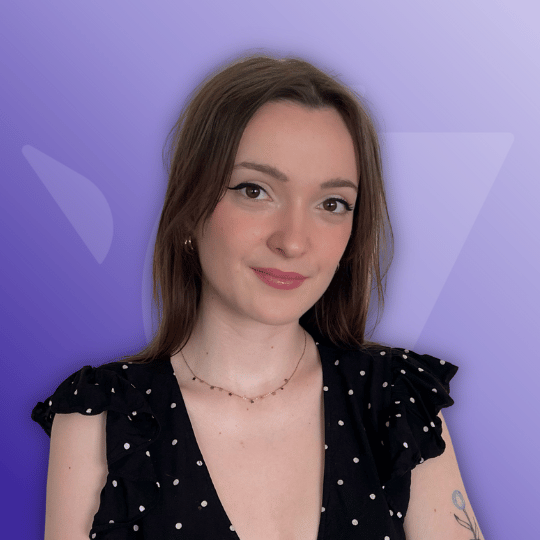 Portrait of a young woman with long brown hair wearing a black polka-dot dress and a delicate necklace, smiling against a purple gradient background.