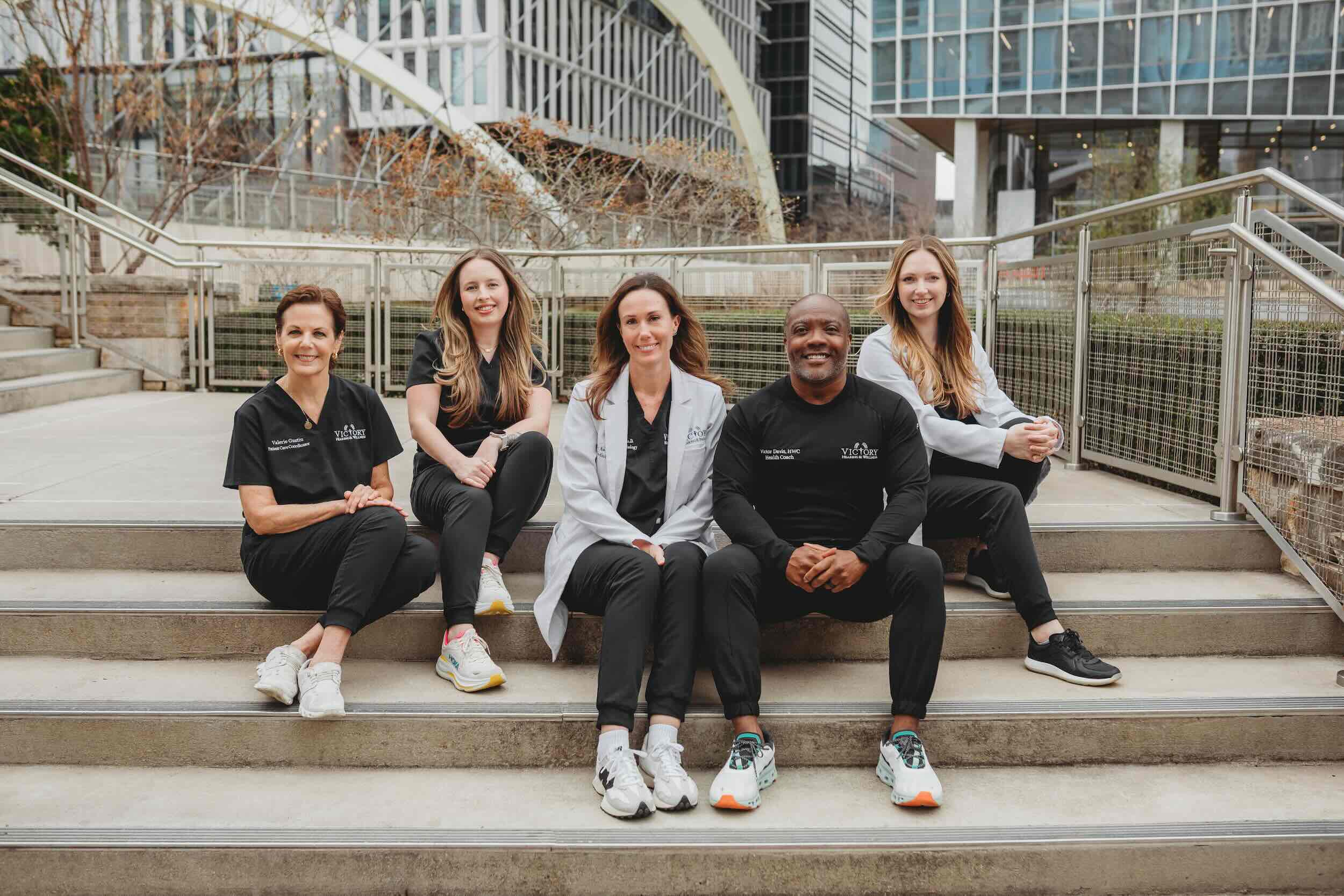 Five healthcare professionals sitting on outdoor steps in black and white Victory Hearing & Balance attire.