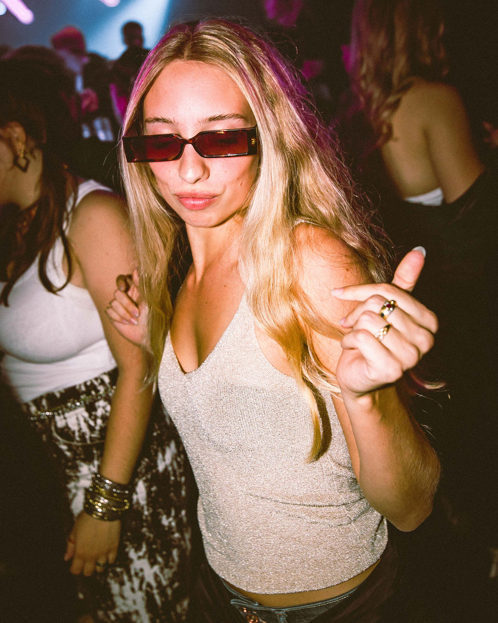 Young woman with long blonde hair wearing red-tinted sunglasses and a sparkling beige tank top dancing at a dimly lit party.