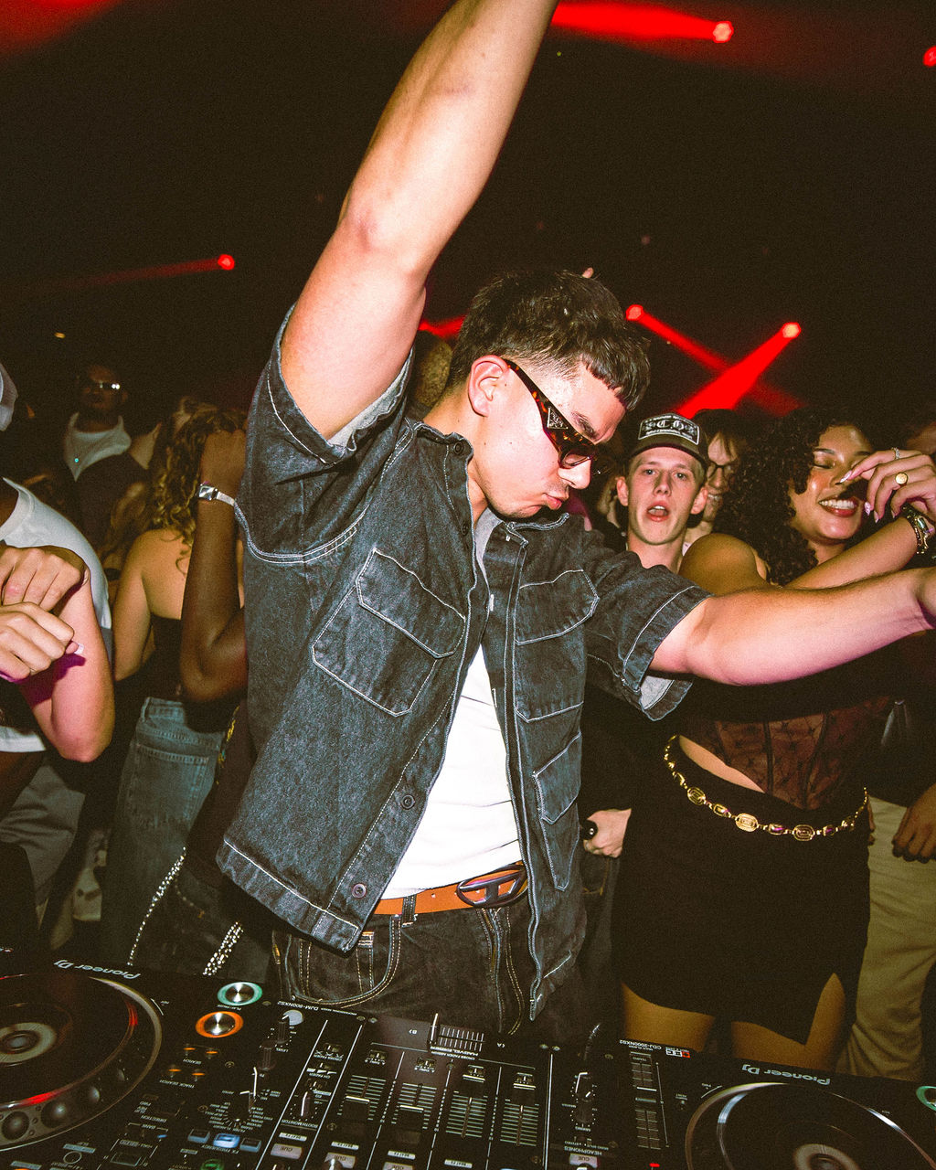 Man wearing sunglasses and denim shirt dancing behind DJ mixer at a crowded nightclub with people enjoying the music.