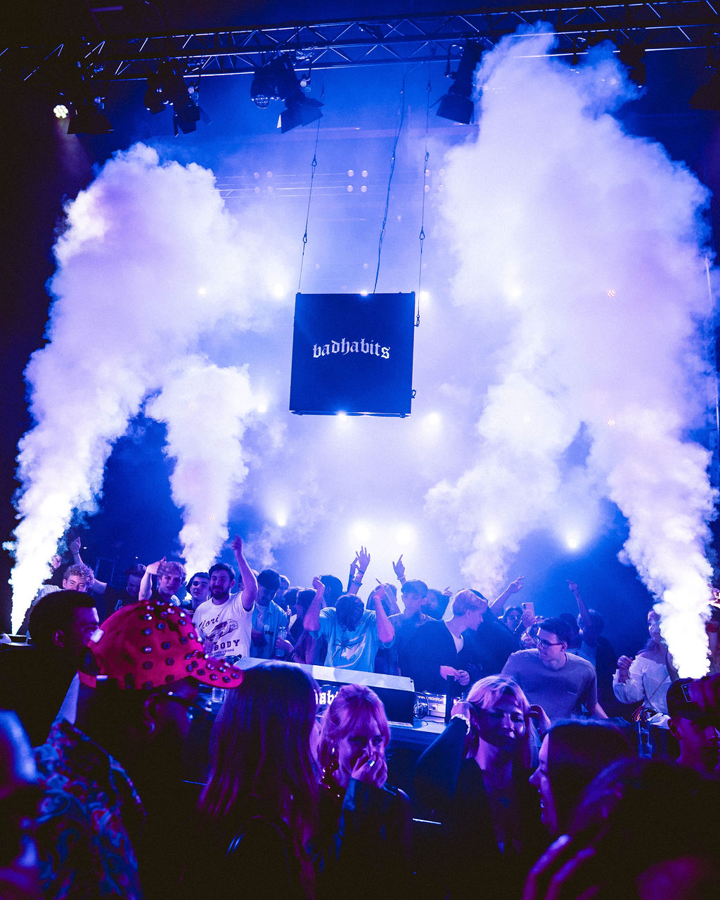 Crowd dancing at a concert with large smoke plumes and a suspended sign reading 'badhabits' in blue lighting.