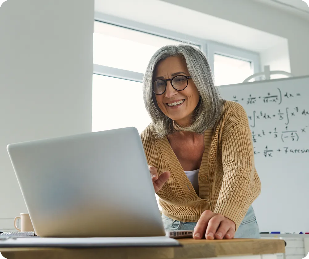 Smiling woman with glasses using a laptop in a bright room with a whiteboard showing mathematical formulas.