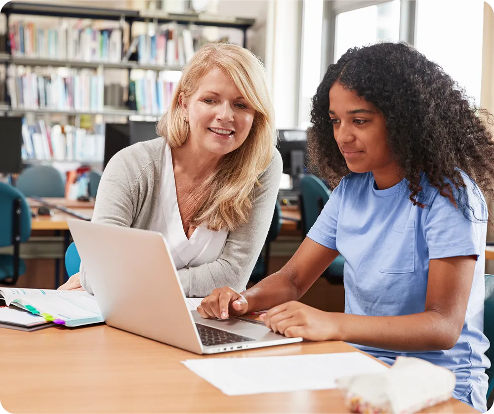 A female teacher assisting a female student with curly hair on a laptop in a classroom.