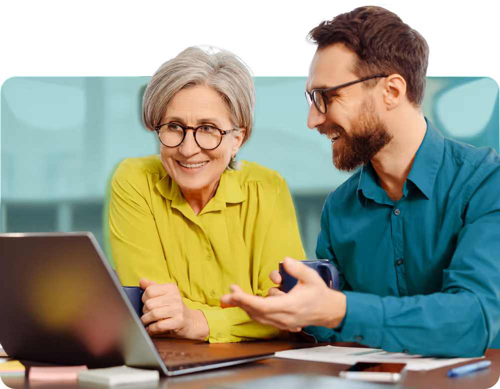 Two colleagues, a woman in a yellow shirt and a man in a teal shirt, smiling and discussing something while looking at a laptop.