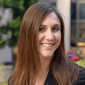 Smiling woman with long brown hair wearing a black blazer, outdoors with blurred greenery and building background.