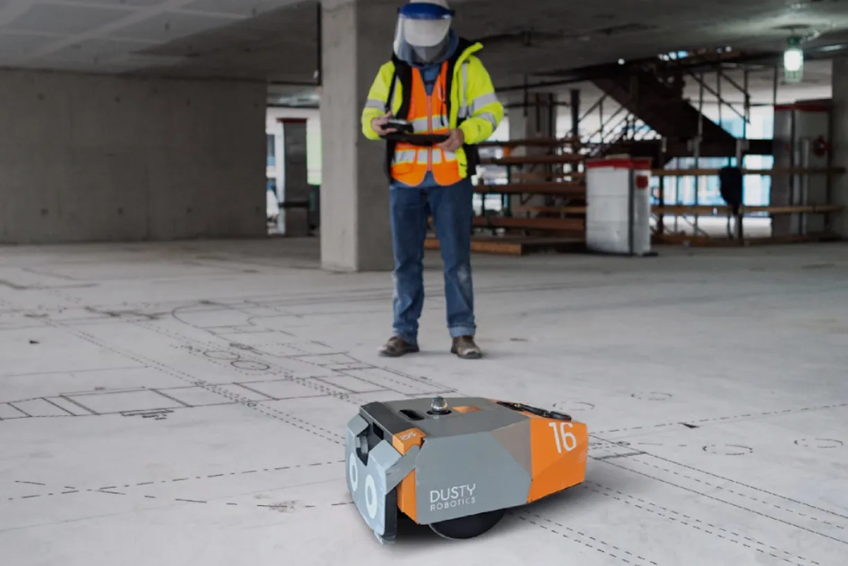 Small orange and gray Dusty Robotics robot on a construction floor with a worker in high-visibility jacket and face shield operating a tablet in the background.