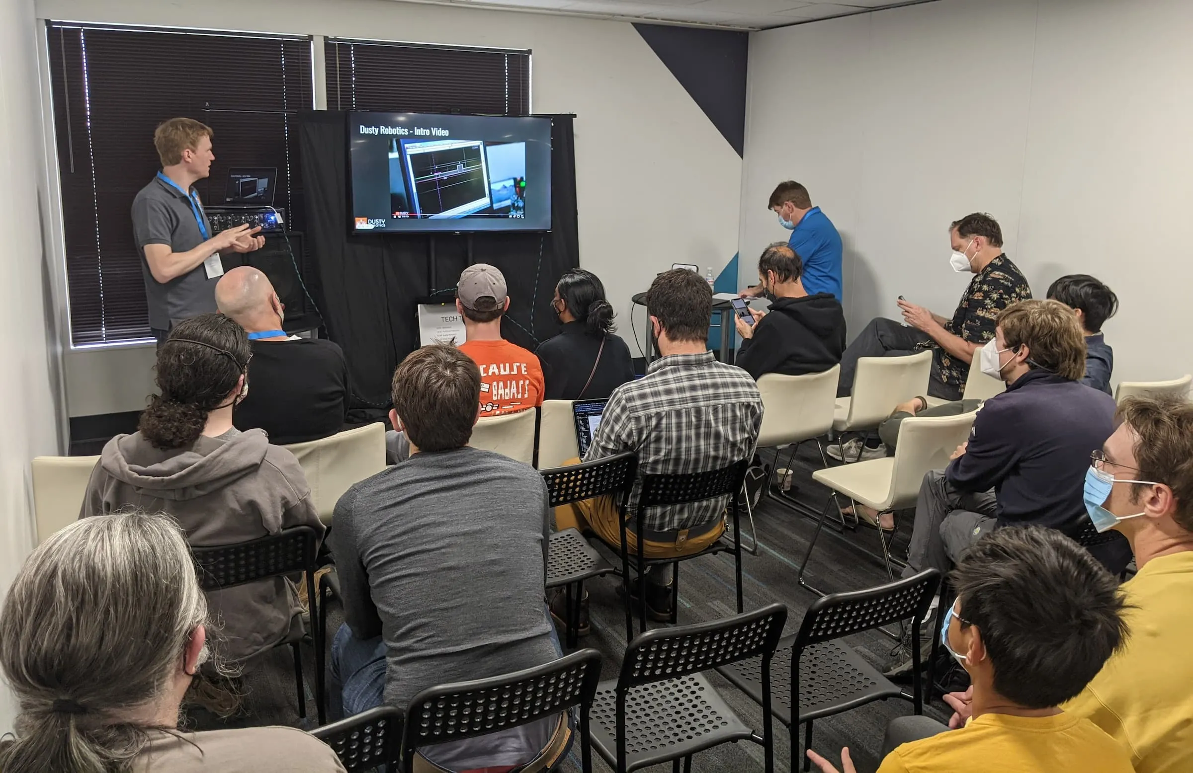 Man giving a technology presentation to an audience seated in a small room, with a video titled 'Dusty Robotics - Intro Video' displayed on a screen.