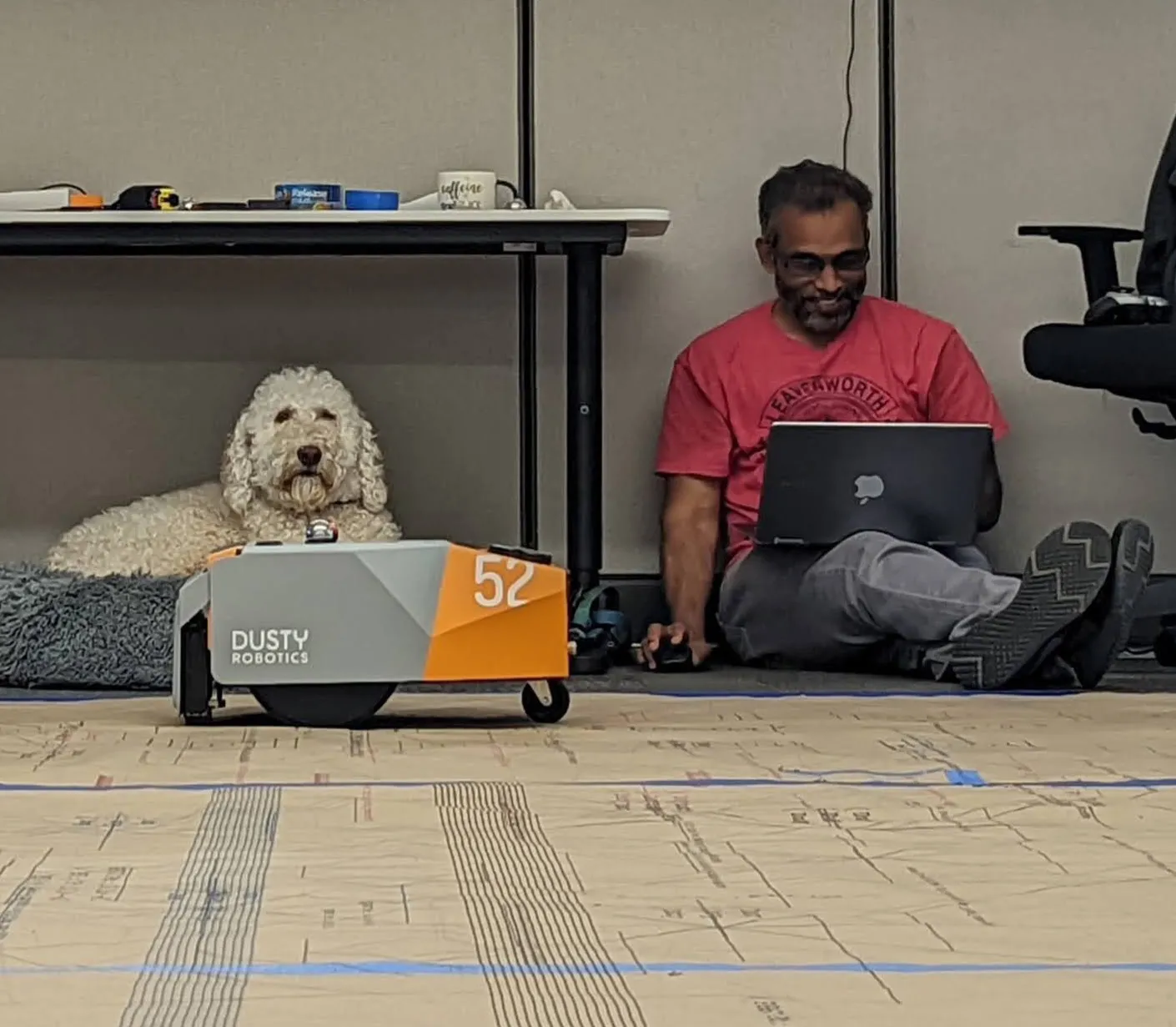 Man sitting on the floor with laptop next to a white curly dog lying on a blanket under a table, with a small robot labeled Dusty Robotics in front.