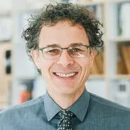 Smiling man with curly hair, glasses, and a gray shirt with a patterned tie, posed in front of a blurred bookshelf.