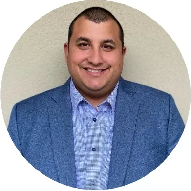 Smiling man with short hair wearing a blue blazer and patterned shirt against a light wall background.