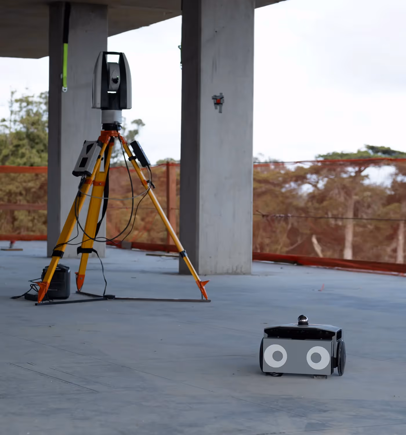 Surveying equipment with a tripod-mounted laser scanner and a small wheeled robot on a construction site floor.
